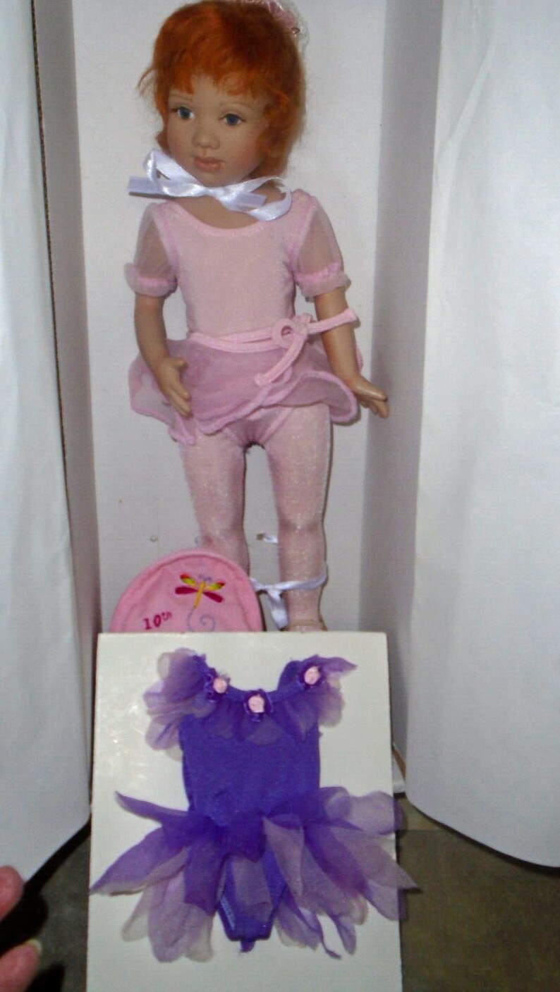 May include: A red-haired doll wearing a pink leotard and tutu with a white ribbon around her neck. The doll is holding a pink and white hat with a dragonfly design and the text "10th" on it. The doll is standing on a white surface with a purple leotard and tutu on a white card.