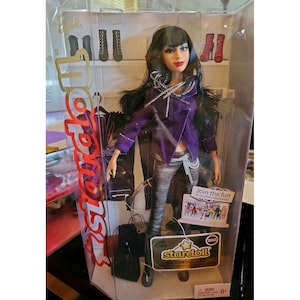 May include: A Stardoll fashion doll in a clear display case. The doll has long black hair, red lipstick, and wears a purple jacket, silver trousers, and black shoes. The packaging features the Stardoll logo and a gift card.
