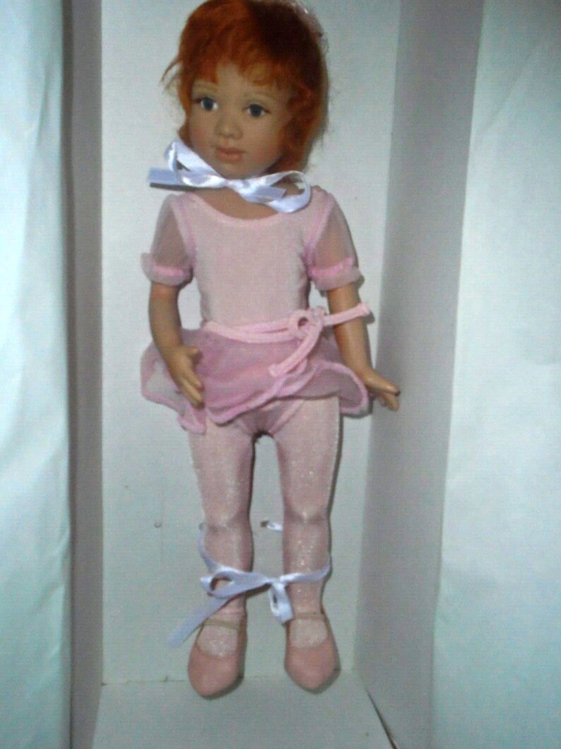 May include: A porcelain doll wearing a pink ballerina costume with white ribbons. The doll has red hair and blue eyes.