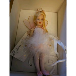 Puede incluir: Muñeca de porcelana con cabello rubio, ojos azules y un disfraz de hada rosa y blanco. La muñeca tiene una falda con plumas, alas rosas y está en una caja con una etiqueta "Robert Tonner".