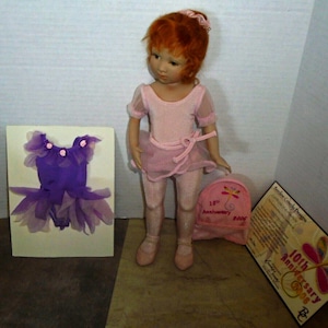 May include: A porcelain doll wearing a pink leotard and tutu, with a purple leotard and tutu on a card behind her. The doll has red hair and is holding a pink backpack with a butterfly design. The backpack has a tag that says "10th Anniversary 2008".