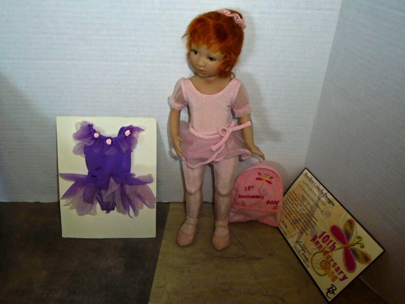 May include: A porcelain doll wearing a pink leotard and tutu, with a purple leotard and tutu on a card behind her. The doll has red hair and is holding a pink backpack with a butterfly design. The backpack has a tag that says "10th Anniversary 2008".
