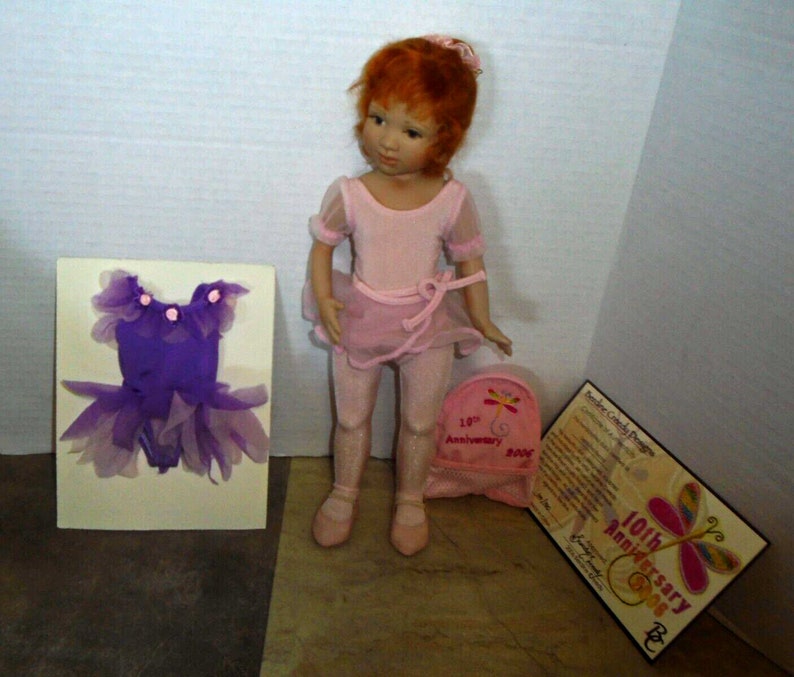 May include: A porcelain doll with red hair wearing a pink leotard and tutu. The doll is holding a pink bag with a butterfly design and the text "10th Anniversary 2006". The doll is also wearing pink ballet shoes. The doll is accompanied by a purple tutu and a certificate.