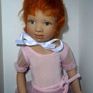 May include: A porcelain doll with red hair wearing a pink leotard and a white ribbon around her neck.