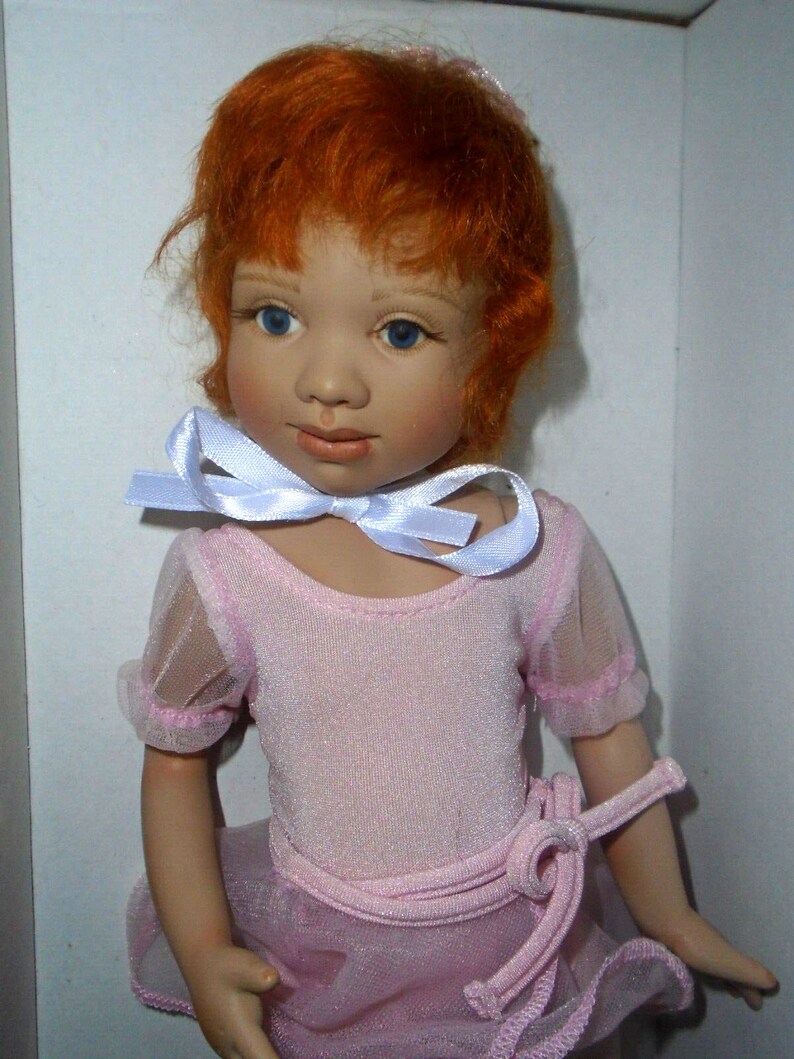May include: A porcelain doll with red hair wearing a pink leotard and a white ribbon around her neck.