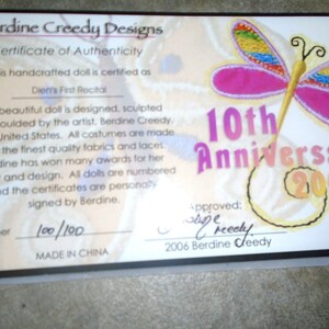 May include: A certificate of authenticity for a handcrafted doll, featuring a pink butterfly with rainbow wings and the text "10th Anniversary 2006". The certificate is numbered "100/100" and states that the doll is designed, sculpted, and moulded by the artist, Berdine Creedy.