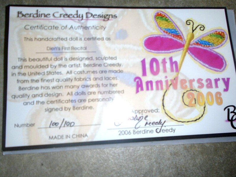 May include: A certificate of authenticity for a handcrafted doll, featuring a pink butterfly with rainbow wings and the text "10th Anniversary 2006". The certificate is numbered "100/100" and states that the doll is designed, sculpted, and moulded by the artist, Berdine Creedy.