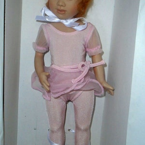 May include: A red-haired doll wearing a pink leotard and tutu with a white ribbon around her neck. She is wearing pink ballet shoes with white ribbons.