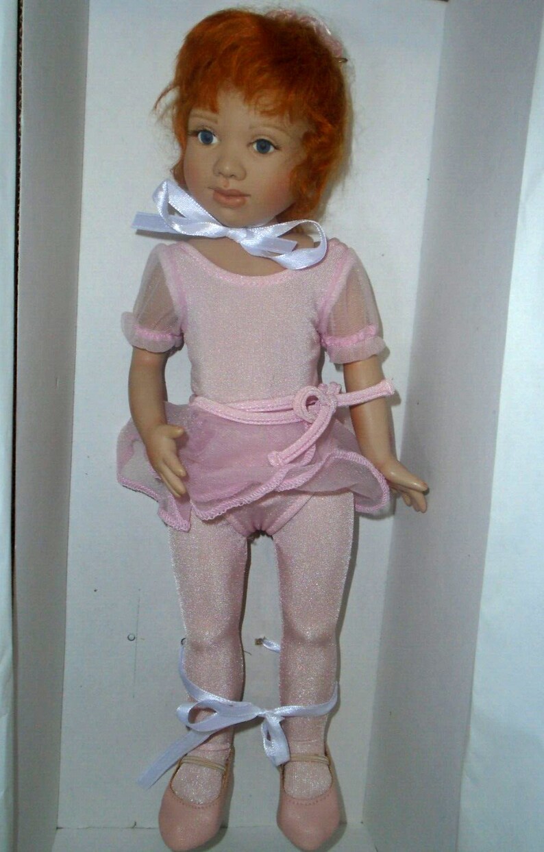 May include: A red-haired doll wearing a pink leotard and tutu with a white ribbon around her neck. She is wearing pink ballet shoes with white ribbons.