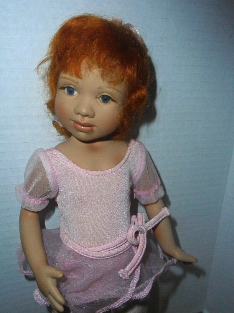 May include: A porcelain doll with red hair wearing a pink leotard and a tulle skirt.