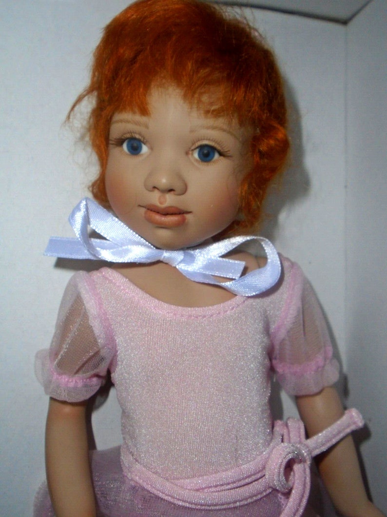 May include: A porcelain doll with red hair wearing a pink leotard and a white ribbon around her neck.