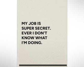 May include: A rectangular, off-white notebook with black text that reads "MY JOB IS SUPER SECRET. EVER I DON'T KNOW WHAT I'M DOING." Below the text are two black horizontal lines and the words "Your Name/Text".