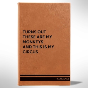 May include: A brown leather-bound journal with the text "TURNS OUT THESE ARE MY MONKEYS AND THIS IS MY CIRCUS" printed in black. A black horizontal bar is at the bottom with space for personalization. The journal has a textured surface.