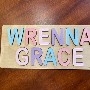 May include: A wooden puzzle with the names "WRENNA GRACE" spelled out in colorful letters. The letters are painted in shades of pink, blue, and purple.