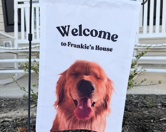 May include: A white rectangular garden flag hanging on a black metal stand. The flag features the black text Welcome to Frankie's House above a photographic print of a golden-colored dog with its eyes closed and tongue hanging out of its mouth.