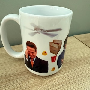 Puede incluir: Taza de cerámica blanca con un collage de imágenes que incluyen un ventilador de techo, un hombre con traje, un vaso rojo, un recipiente para llevar marrón y patitos de goma. La taza tiene un asa curva y un acabado brillante.