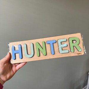 May include: A wooden puzzle with the name "HUNTER" spelled out in colorful letters. The letters are in shades of blue, green, and light blue. The puzzle is made by "PuzzleCo."