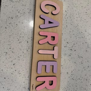 May include: A wooden puzzle with the name "CARTER" spelled out in pink and purple letters. The letters are cut out and fit into the wooden board. The board is light brown and has the text "Figs & Co" printed on the bottom left corner.