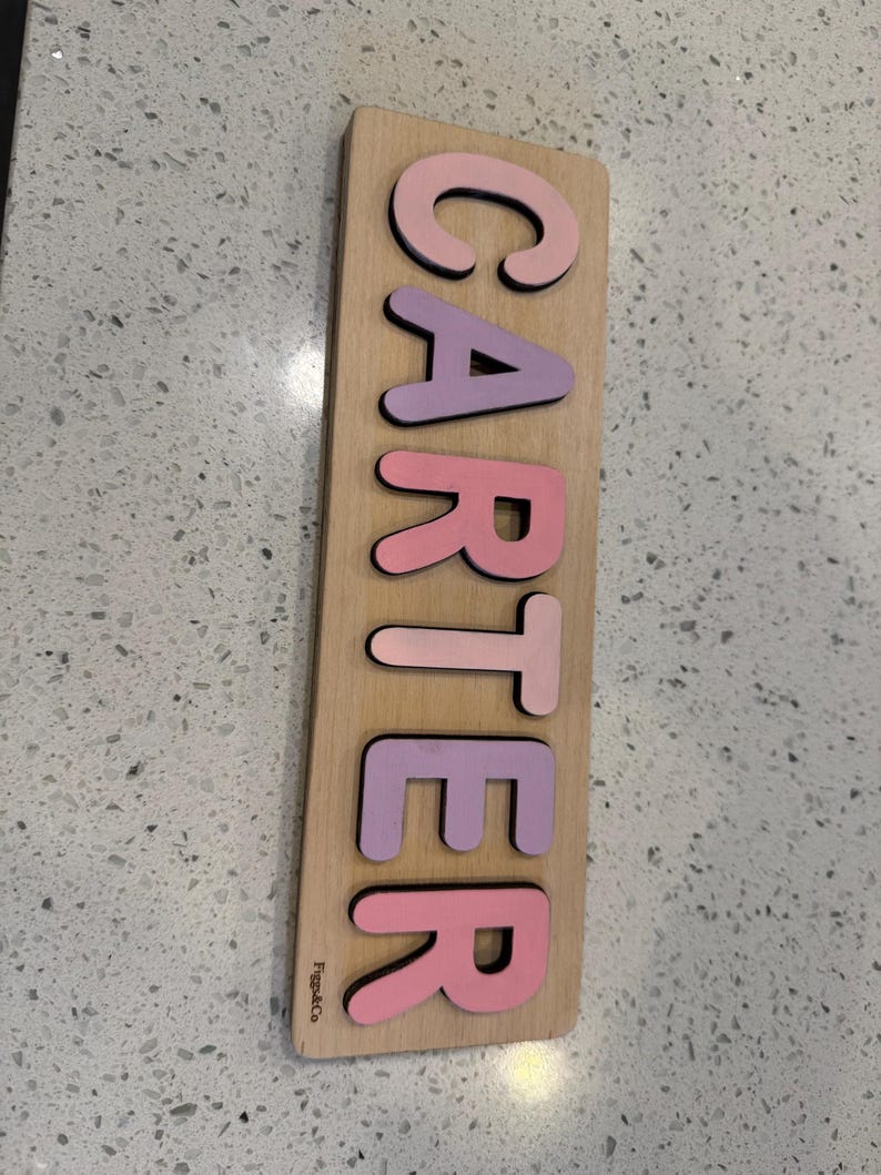 May include: A wooden puzzle with the name "CARTER" spelled out in pink and purple letters. The letters are cut out and fit into the wooden board. The board is light brown and has the text "Figs & Co" printed on the bottom left corner.
