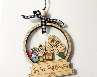 May include: A wooden snow globe ornament with a clear plastic dome. Inside the dome are various wooden cutouts, including a calendar with the date June 24, a measuring tape with the measurement 20 inches, a clock with the time 9:35 PM, a small tree, a snowflake, a star, a butterfly, and a heart. The base of the ornament is engraved with the text "Sayfori's First Christmas 2023".