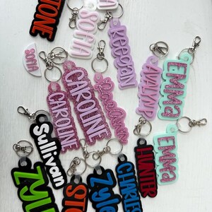May include: A collection of personalized keychains in various colors. Each keychain features a name, such as "Stone", "Sofia", and "Caroline", in bold, contrasting colors. The keychains have a glossy finish and are attached to silver key rings.