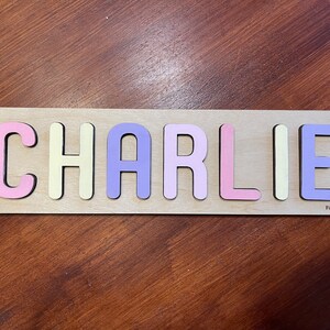 May include: A wooden puzzle with the name "CHARLIE" spelled out in colorful letters. The letters are pink, yellow, purple, and light pink. The puzzle is on a wooden surface.
