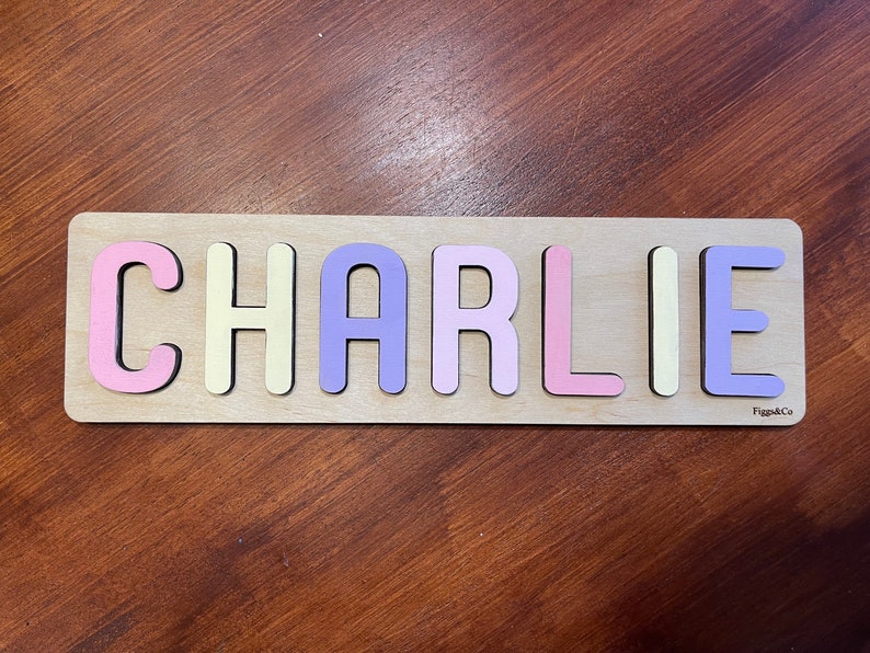 May include: A wooden puzzle with the name "CHARLIE" spelled out in colorful letters. The letters are pink, yellow, purple, and light pink. The puzzle is on a wooden surface.