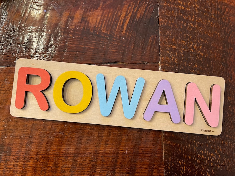 May include: A wooden name puzzle with the name "ROWAN" spelled out in colorful letters. The letters are red, yellow, blue, purple, and pink. The puzzle is on a wooden surface.