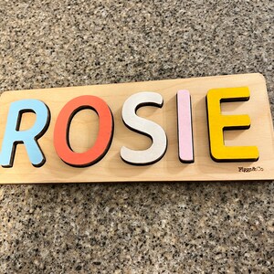 May include: A wooden puzzle with the name "ROSIE" spelled out in colorful letters. The letters are blue, red, white, pink, and yellow. The puzzle is made by "Figgs & Co."