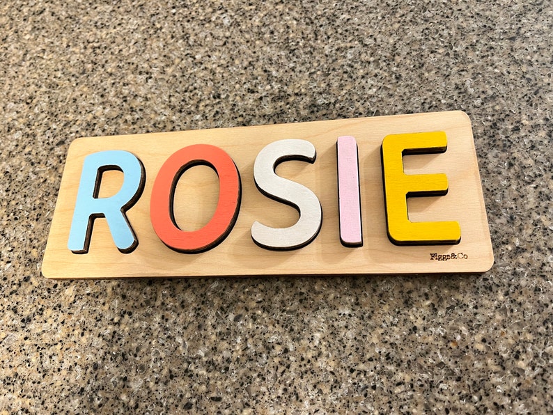 May include: A wooden puzzle with the name "ROSIE" spelled out in colorful letters. The letters are blue, red, white, pink, and yellow. The puzzle is made by "Figgs & Co."