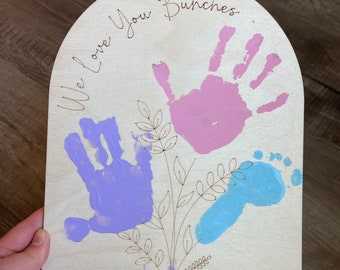 May include: Wooden plaque with an arched top, featuring handprints and a footprint in pink, purple, and blue, arranged as a bouquet. The text "We Love You Bunches" and "Happy Mothers Day" are engraved, along with names and the year 2024.