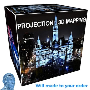 May include: A cube-shaped display featuring the text PROJECTION 3D MAPPING on the top edge. The front face shows a night scene of a large building illuminated with a projection, while the side face displays a blue and red abstract light pattern.