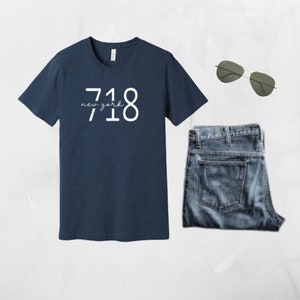 May include: A navy blue t-shirt with the text "718 new york" printed in white on the front. The t-shirt is paired with blue jeans and sunglasses.