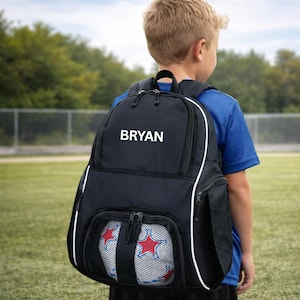 May include: Black backpack with the name "BRYAN" in white. The backpack has white trim and a front pocket holding a soccer ball. The image is taken on a green field.