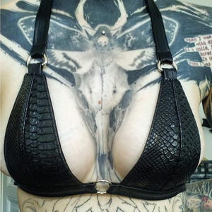 Pode incluir: Um sutiã preto com padrão de pele de cobra, estilo halter. O sutiã apresenta copas triangulares e detalhes de anéis de metal no centro e nas fixações das alças. As alças são finas e pretas.