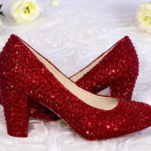 May include: A pair of red glitter-covered high heels with a block heel. The shoes have a rounded toe and a neutral-colored interior. The background features white flowers and a textured white surface.