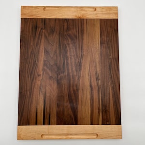 May include: A large rectangular cutting board made of walnut wood with a maple wood border and handles. The board has a natural wood finish.