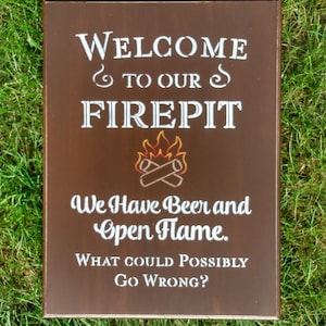 May include: A brown wooden sign with white text that reads "Welcome to our firepit. We have beer and open flame. What could possibly go wrong?" The sign has a stylized image of a campfire with crossed logs.