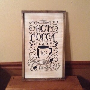 May include: A rectangular wooden sign with a distressed white finish and a dark brown frame. The sign features black text that reads OLD-FASHIONED HOT COCOA, MARSHMALLOWS, and WHIPPED CREAM, along with a graphic of a mug labelled 10 cents.
