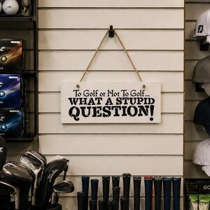May include: A rectangular white wooden sign hanging from a jute rope, featuring the black printed text: To Golf or Not To Golf... WHAT A STUPID QUESTION! The sign is displayed on a slatted wall alongside various golf equipment including hats and clubs.