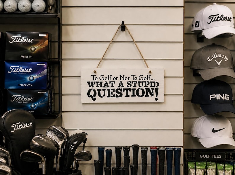 May include: A rectangular white wooden sign hanging from a jute rope, featuring the black printed text: To Golf or Not To Golf... WHAT A STUPID QUESTION! The sign is displayed on a slatted wall alongside various golf equipment including hats and clubs.