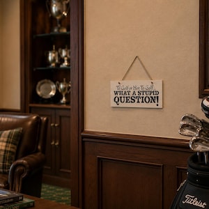 May include: A rectangular white wooden sign hanging by a jute rope, featuring black text that reads: To Golf or Not To Golf... WHAT A STUPID QUESTION! The sign is mounted on a light-colored wall above dark wood paneling in a room with golf decor.