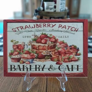 May include: A red and white sign with the text "Strawberry Patch" and "Bakery & Cafe". The sign features a painting of a strawberry cake with strawberries and cupcakes.