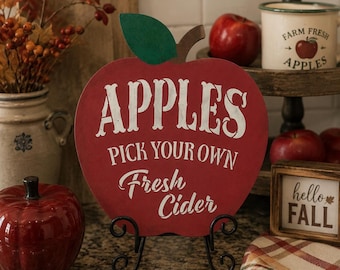 May include: A red apple-shaped wooden sign featuring a green leaf and white text that reads "APPLES PICK YOUR OWN Fresh Cider." The sign is supported by a black metal scrollwork stand, creating a decorative display piece for seasonal decor.