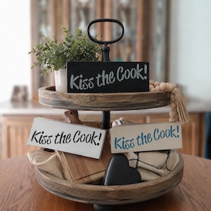 May include: A two-tiered wooden tray featuring three rectangular signs with the text Kiss the Cook! in different color combinations, a small potted green plant, a wooden bead garland with a tassel, a wooden cutting board, and a black heart.
