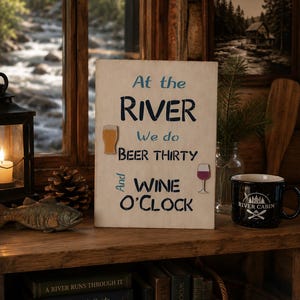 May include: A rectangular white wooden sign with dark blue and light blue text that reads: At the RIVER We do BEER THIRTY And WINE O'CLOCK. The sign features a small wooden cutout of a beer glass and a wine glass attached to the front surface.