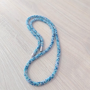 Può includere: Una collana di perline nei toni del blu, bianco e argento. La collana è realizzata con piccole perline, creando un aspetto strutturato simile a una corda. La chiusura è color argento. La collana è esposta su una superficie di legno chiaro.