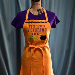 Puede incluir: Delantal naranja con el texto "IT'S FUN STIRRING THE POT" y un gráfico de caldero. El delantal tiene un borde morado y blanco estampado con fantasmas y calabazas. Tiene dos bolsillos y una cinta para la cintura.