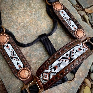May include: A brown leather horse halter with decorative beadwork in white, black, and orange. The halter features copper-colored conchos and a black nylon rope and straps. The design is intricate and detailed.