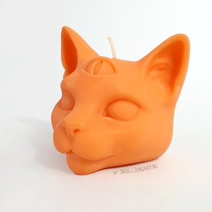 Puede incluir: Vela naranja con forma de cabeza de gato con un tercer ojo y la boca cerrada. La vela est&aacute; hecha de cera y tiene una mecha de algod&oacute;n. La vela est&aacute; sobre un fondo blanco. Por Skulldinghot.nl